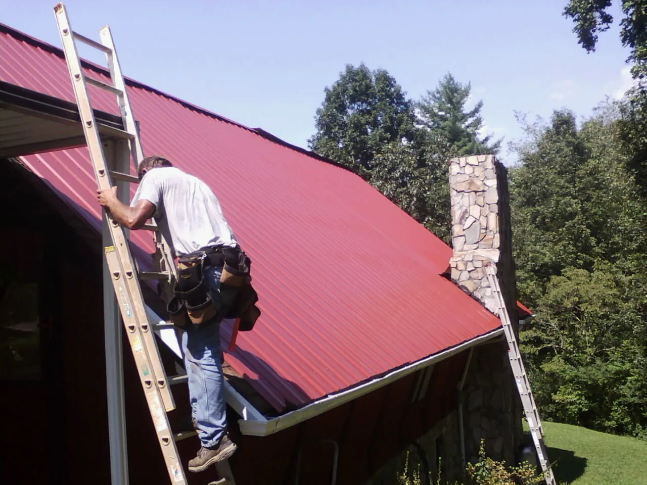 Roofer on a ladder beside a red metal roof and stone chimney — Soffit & Fascia Repair by Roofing Brothers in Cygnet